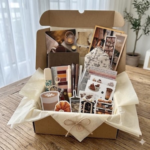 Könnte beinhalten: Eine Geschenkschachtel aus Karton, gefüllt mit Schreibwaren wie einem braunen Notizbuch, Stiften, einem Lineal, einem gemusterten Stoffbeutel und verschiedenen Aufklebern mit Kaffee-, Gebäck- und Buchmotiven, ausgelegt mit cremefarbenem Seidenpapier.