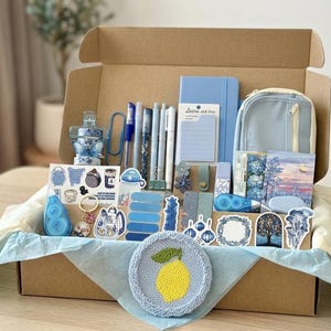 Könnte beinhalten: Eine offene Geschenkbox mit blauen Schreibwaren, darunter Stifte, ein Notizbuch und Aufkleber. Ein gestrickter Untersetzer mit gelbem Zitronenmuster befindet sich am Boden. Die Box enthält ein Federmäppchen und weiteres Zubehör.
