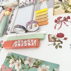 May include: A collection of stationery items with a floral theme. Includes notebooks, sticky notes, washi tape, highlighters, and decorative stickers. The color palette features soft pastels, with accents of pink, green, and yellow. The items are arranged on a white surface.