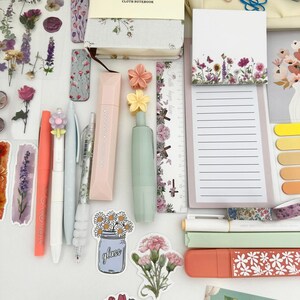 May include: An assortment of stationery items, including pens, stickers, a notebook, and a notepad, arranged on a white surface. The items feature floral designs and pastel colors, with the text "FLOWER & TREE CLOTH NOTEBOOK" visible on the notebook.