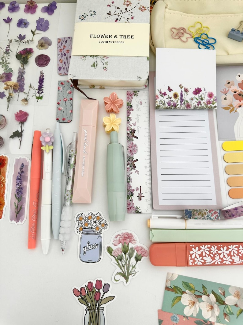 May include: An assortment of stationery items, including pens, stickers, a notebook, and a notepad, arranged on a white surface. The items feature floral designs and pastel colors, with the text "FLOWER & TREE CLOTH NOTEBOOK" visible on the notebook.