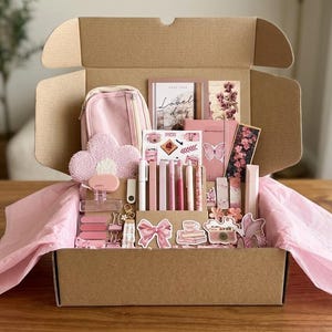 Könnte beinhalten: Ein Karton mit rosa Schreibwaren und Accessoires. Enthalten sind ein Federmäppchen, Notizbücher, Stifte und Aufkleber. Die Box ist mit rosa Seidenpapier ausgekleidet und steht auf einer Holzoberfläche.