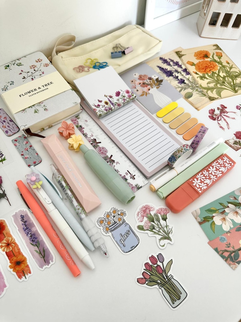 May include: An assortment of stationery items, including pens, notebooks, stickers, and sticky notes, arranged on a white surface. The items feature floral designs and pastel colors, creating a cohesive aesthetic. A pencil case and decorative paper are also visible.