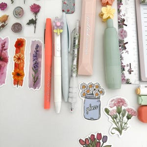 May include: An assortment of stationery items, including pens, correction tape, and stickers. The pens are in coral, white, and light blue. The correction tape is pink. Stickers feature flowers and the text "glass".