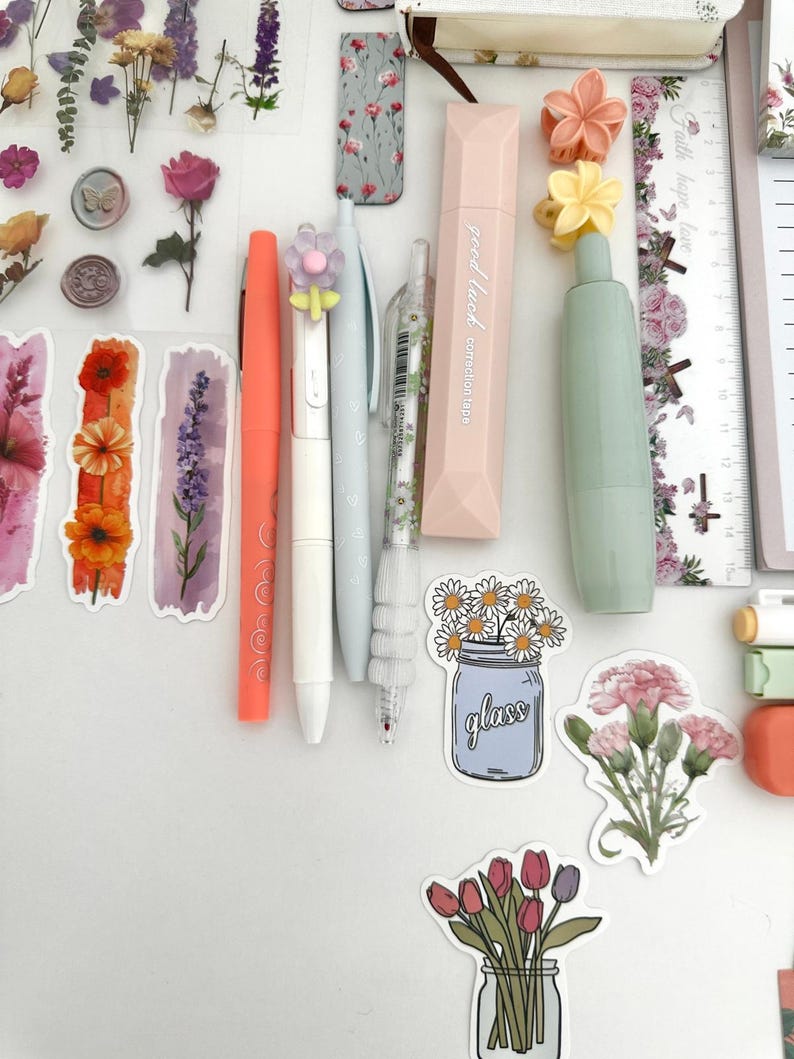 May include: An assortment of stationery items, including pens, correction tape, and stickers. The pens are in coral, white, and light blue. The correction tape is pink. Stickers feature flowers and the text "glass".