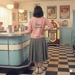 Op de afbeelding: Een vrouw in een roze jasje met "Pink Ladies" op de rug staat in een retro diner. Ze draagt een grijze rok en witte sokken met roze sandalen. De diner heeft een jukebox, posters en een zwart-wit geblokte vloer.