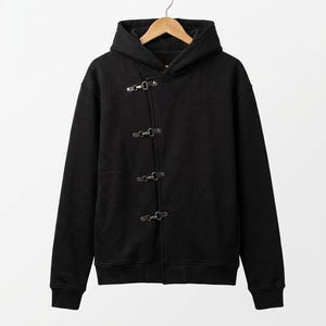 May include: Black hooded sweatshirt with a toggle closure. The sweatshirt is hanging on a wooden hanger. The closure has four metal toggle clasps. The fabric appears to be a medium-weight material.
