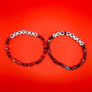May include: Two beaded bracelets featuring white letter beads. The left bracelet spells MCQUEEN with red, black, and clear beads. The right bracelet spells TOW MATER with dark brown, black, and clear beads, plus two small bright blue beads.