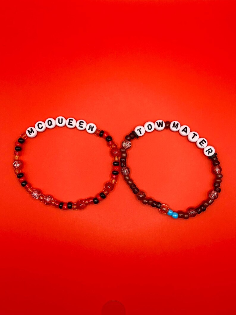 May include: Two beaded bracelets featuring white letter beads. The left bracelet spells MCQUEEN with red, black, and clear beads. The right bracelet spells TOW MATER with dark brown, black, and clear beads, plus two small bright blue beads.