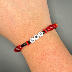 Op de afbeelding: Een kralenarmband met rode en zwarte kralen. De armband heeft witte letterkralen met de letters "K G" met een hartsymbool. De armband wordt om een pols gedragen.