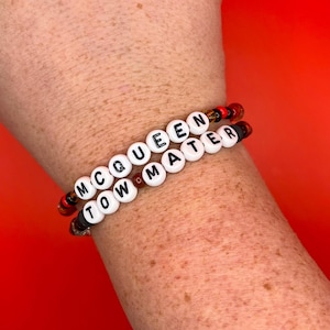 May include: Two elastic bracelets featuring white round beads with black letters spelling out MCQUEEN and TOW MATER. The bracelets are accented with smaller, translucent red, orange, and black beads interspersed between the lettered beads.