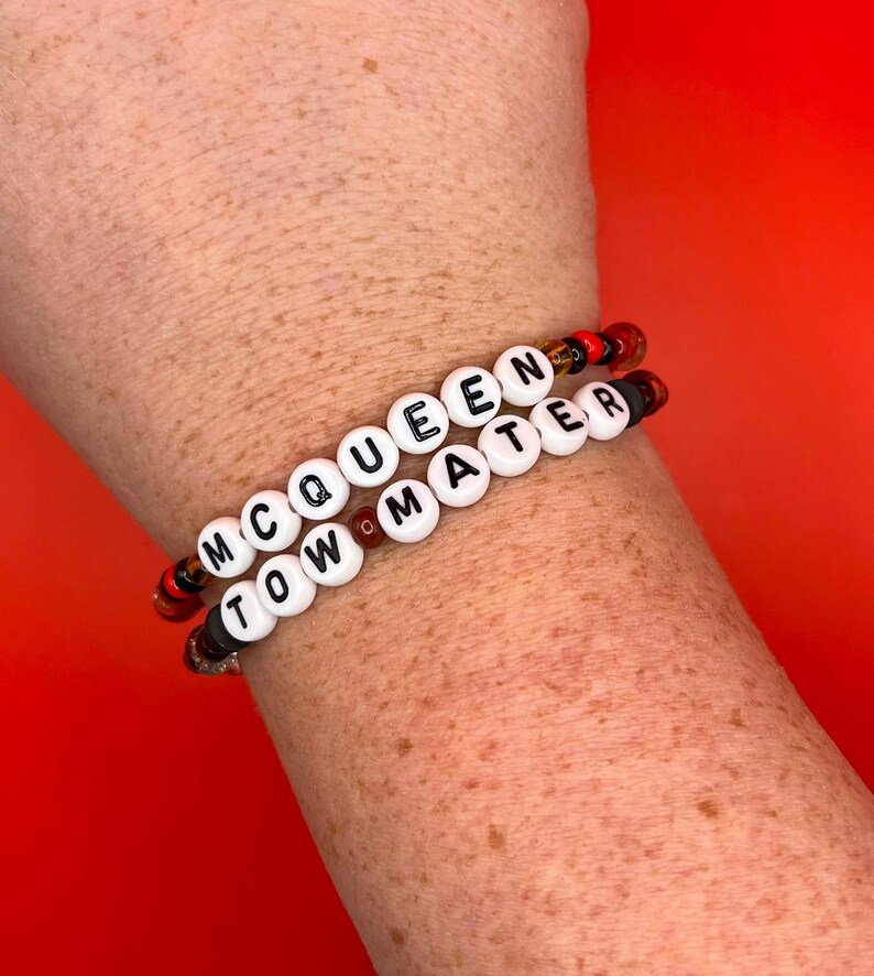 May include: Two elastic bracelets featuring white round beads with black letters spelling out MCQUEEN and TOW MATER. The bracelets are accented with smaller, translucent red, orange, and black beads interspersed between the lettered beads.