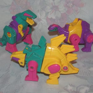 May include: Three brightly coloured toy dinosaurs in green, yellow, purple, and pink. The toys have articulated limbs and a retro design. One dinosaur has a spiky back, while another has a long neck. The toys are likely from the 1980s or 1990s.