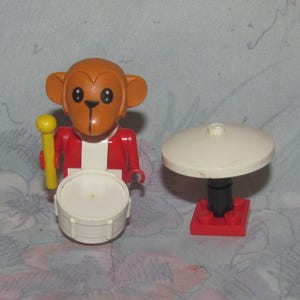 May include: A toy monkey drummer figure with a red jacket, white shirt, and a white drum. The monkey holds a yellow drumstick and is next to a white cymbal on a red base. A fun toy for kids.