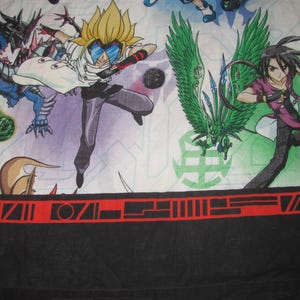 Peut inclure: Un drap en tissu avec un imprimé anime coloré représentant des personnages et des dragons sur fond blanc. La partie inférieure présente une bordure noire unie avec une bande rouge horizontale ornée de symboles géométriques noirs.