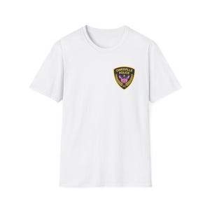 Op de afbeelding: Wit T-shirt met een schouderpatch met de tekst "Oinksville Police" boven een roze varkensgezicht. De patch is omlijst door een gouden rand en tarwearen. Het shirt heeft korte mouwen en een ronde hals.