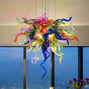 May include: A colourful glass chandelier with an artistic design. It features swirling, elongated glass elements in red, orange, yellow, green, blue, and purple. The chandelier hangs from thin wires, creating a floating effect. A statement piece for any room.