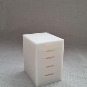 May include: A small, matte white storage unit featuring a rectangular frame and four stacked drawers. Each drawer is equipped with a horizontal, recessed handle slot centered on the front panel, providing a clean and uniform appearance.