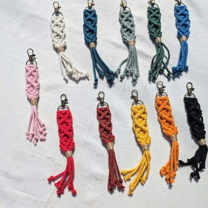 Peut inclure: Une collection de porte-clés en macramé de différentes couleurs : rose, blanc, sarcelle, vert, bleu, rouge, marron, jaune, orange et noir. Chaque porte-clés présente un motif tissé avec un pompon et un fermoir doré.