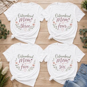 Pode incluir: Quatro camisetas brancas de manga curta com gráficos florais e texto. As camisetas exibem as frases Outnumbered Mom of three, Outnumbered Mom of four, Outnumbered Mom of five e Outnumbered Mom of six em tinta roxa e verde.