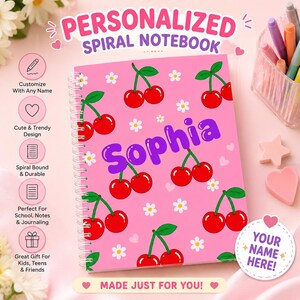 Könnte beinhalten: Ein pinkfarbenes Spiralnotizbuch mit einem wiederkehrenden Muster aus roten Kirschen und weißen Blumen. Der Name Sophia ist in großen lila Buchstaben in der Mitte aufgedruckt. Das Cover enthält den Text Personalized Spiral Notebook.