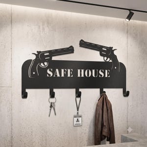 Puede incluir: Un perchero de pared de metal negro con el texto SAFE HOUSE recortado y dos siluetas de revólver en la parte superior. El soporte cuenta con cinco ganchos, de los cuales dos sostienen un llavero y una tarjeta de identificación.