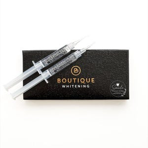 Può includere: Due siringhe in plastica trasparente riempite di gel sbiancante sono posizionate sopra una scatola rettangolare nera. La scatola presenta un motivo floreale in rilievo, il testo dorato BOUTIQUE WHITENING e un logo circolare con un'icona a dente.