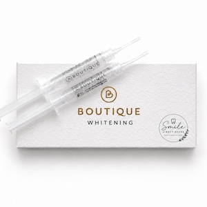 Op de afbeelding: Twee doorzichtige plastic spuiten gevuld met gel, gelabeld met Boutique 6% Hydrogen Peroxide, liggen op een rechthoekige witte doos. De doos bevat de gouden tekst Boutique Whitening en een cirkelvormig logo met Smile Direct Store.
