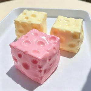 May include: Three cube-shaped objects with a smooth, glossy finish and circular indentations across their surfaces. One cube is light pink, while the other two are pale yellow. The cubes are arranged in a cluster on a white surface.