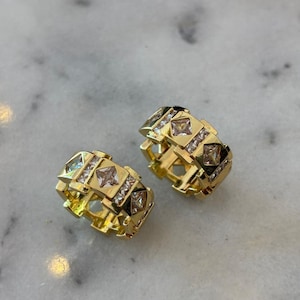 Puede incluir: Dos anillos dorados con un diseño geométrico. Cada anillo presenta piedras claras cuadradas y rectangulares. Los anillos están sobre una superficie de mármol.