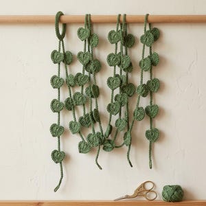 Puede incluir: Una guirnalda de enredadera tejida a ganchillo en color verde salvia con múltiples hojas en forma de corazón unidas a largos hilos. La guirnalda cuelga de una varilla de madera, con un pequeño ovillo de lana verde a juego y tijeras doradas.