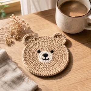 May include: A round, tan crochet coaster shaped like a bear face. The coaster features two small rounded ears at the top, a circular cream-colored muzzle, and embroidered black yarn details for the eyes, nose, and a small curved mouth.