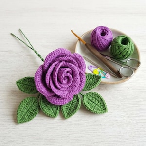 May include: A crocheted purple rose with green leaves on a stem, accompanied by a small ceramic dish containing a ball of purple yarn, a ball of green yarn, a brown crochet hook, a pair of silver scissors, and two colorful plastic stitch markers.