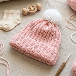 Op de afbeelding: Een lichtroze gebreide muts met een dikke geribbelde textuur en een grote witte wollen pompon aan de bovenkant. De muts heeft een omgeslagen boord en ligt naast een paar crèmekleurige sokken, een houten rammelaar en een haaknaald.