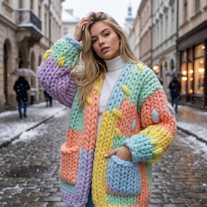 Puede incluir: Un cárdigan de punto grueso con un patrón de bloques de color en tonos amarillo pastel, azul claro, melocotón suave y lavanda. La prenda tiene mangas largas, una tapeta frontal acanalada y dos bolsillos, sobre un suéter de cuello alto blanco.