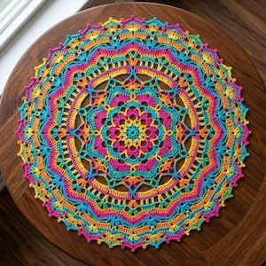 May include: A circular crochet doily featuring a vibrant, multi-coloured mandala pattern. The design incorporates concentric rings of bright yellow, hot pink, turquoise, and orange yarn, arranged in a complex, symmetrical, and intricate lace-like structure.