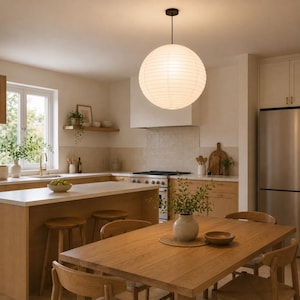 May include: A well-lit kitchen with light wood cabinetry, a white countertop, and a stainless steel refrigerator. A large, round paper lantern hangs above a wooden dining table with chairs. A vase of greenery sits on the table.