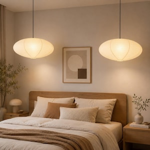 Puede incluir: Dos lámparas colgantes ovaladas con pantallas de papel iluminan una habitación. La cama es de madera, con ropa de cama en tonos neutros. Un cuadro enmarcado cuelga de la pared. Las lámparas emiten una luz cálida y tenue.