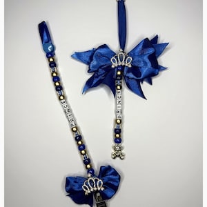 May include: Two blue ribbon accessories featuring gold and blue beads, silver crown charms, and white letter beads spelling PRINCE. One accessory includes a silver teddy bear charm and a metal clip, while the other features a large blue bow and clip.