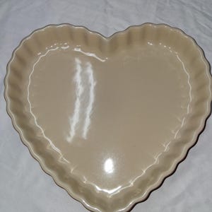 May include: A heart-shaped baking dish with a smooth, glossy, light beige finish. The dish features a scalloped, fluted edge design around the entire perimeter, creating a decorative border for the interior basin of the ceramic bakeware piece.