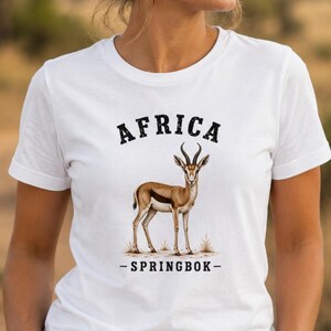 May include: A white short-sleeved t-shirt featuring a graphic illustration of a brown and white springbok standing in grass. The text "AFRICA" is printed in black above the animal, and the text "- SPRINGBOK -" is printed in black below it.