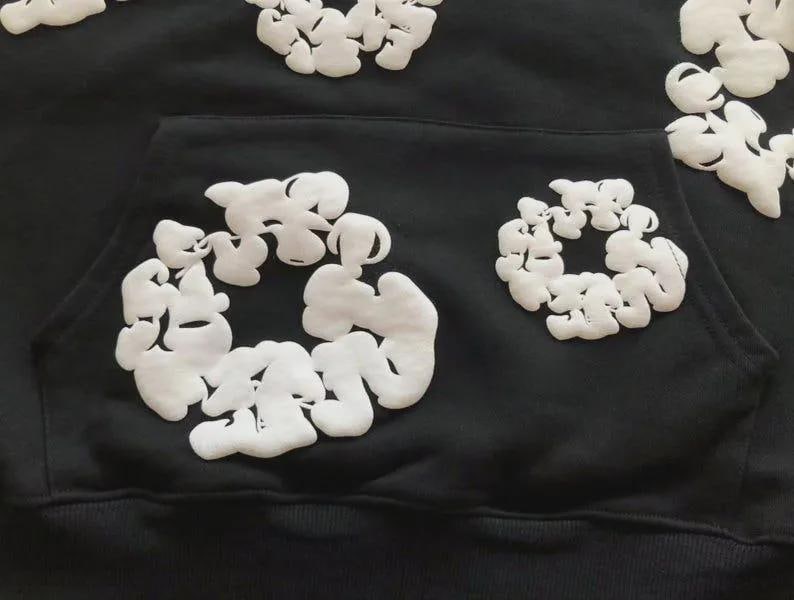 May include: Black hoodie with white, puffy, abstract floral designs. The hoodie has a front pocket and a drawstring hood. The white designs are clustered in circular patterns across the front and sleeves, creating a unique, textured look.