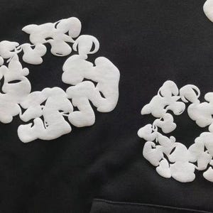 May include: A close-up shot of a black garment with white, cloud-shaped embroidery. The designs are arranged in circular patterns and have a textured finish. The dark fabric provides a strong contrast to the white embroidery.