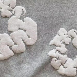 May include: A close-up shot of grey fabric adorned with white, cloud-like appliqu&eacute;s. The appliqu&eacute;s have a textured finish, contrasting with the smooth background. The design is simple and contemporary, with a modern aesthetic.