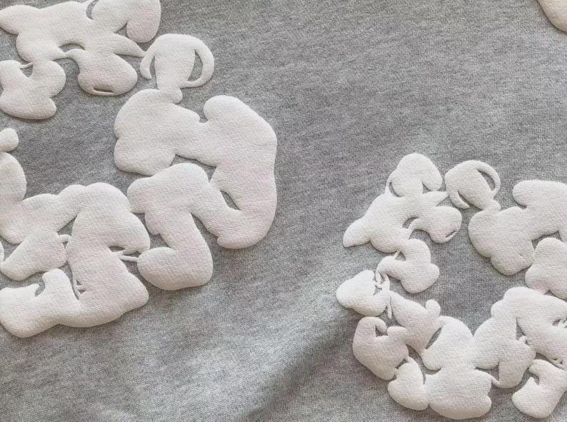 May include: A close-up shot of grey fabric adorned with white, cloud-like appliqu&eacute;s. The appliqu&eacute;s have a textured finish, contrasting with the smooth background. The design is simple and contemporary, with a modern aesthetic.