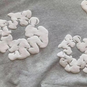 May include: A grey sweatshirt featuring white cloud-shaped appliqu&eacute;s in circular arrangements. The fabric's texture and the design suggest a casual, comfortable garment, suitable for everyday wear.