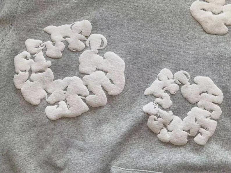 May include: A grey sweatshirt featuring white cloud-shaped appliqu&eacute;s in circular arrangements. The fabric's texture and the design suggest a casual, comfortable garment, suitable for everyday wear.
