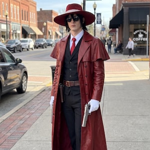 May include: A dark red leather trench coat with a capelet, worn over a black vest, black trousers, and a white dress shirt with a red tie. The outfit includes a wide-brimmed dark red hat, black sunglasses, white gloves, and a brown leather belt.