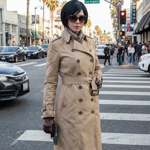 May include: A tan trench coat with a belt and button details. The coat is worn over a scarf and sunglasses. The person is standing on a crosswalk in Los Angeles, holding a handgun.