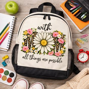 Peut inclure: Un sac à dos crème avec une base et des bretelles noires. Le devant présente le mot "MOM" avec un motif de marguerite et de fleurs, des accents d'imprimé léopard et la phrase "all things are possible". Une pomme verte, des fournitures scolaires et un réveil rouge sont visibles.